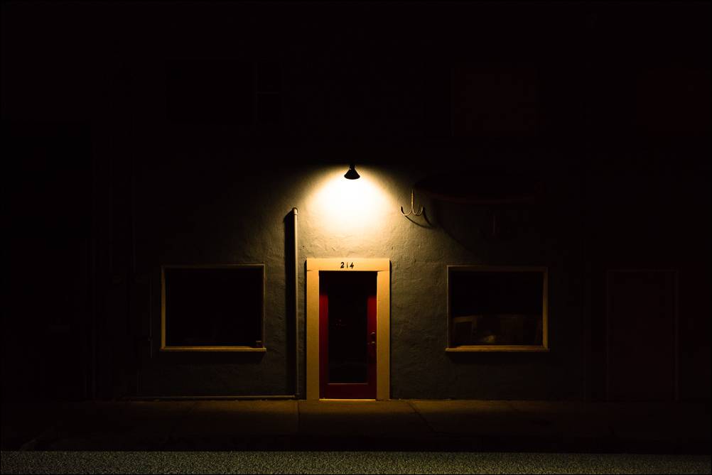 Explore. Behind the Red Door. A photograph by Jeff Kauffman of an olive-green storefront with a red door at night under dramatic street lighting.